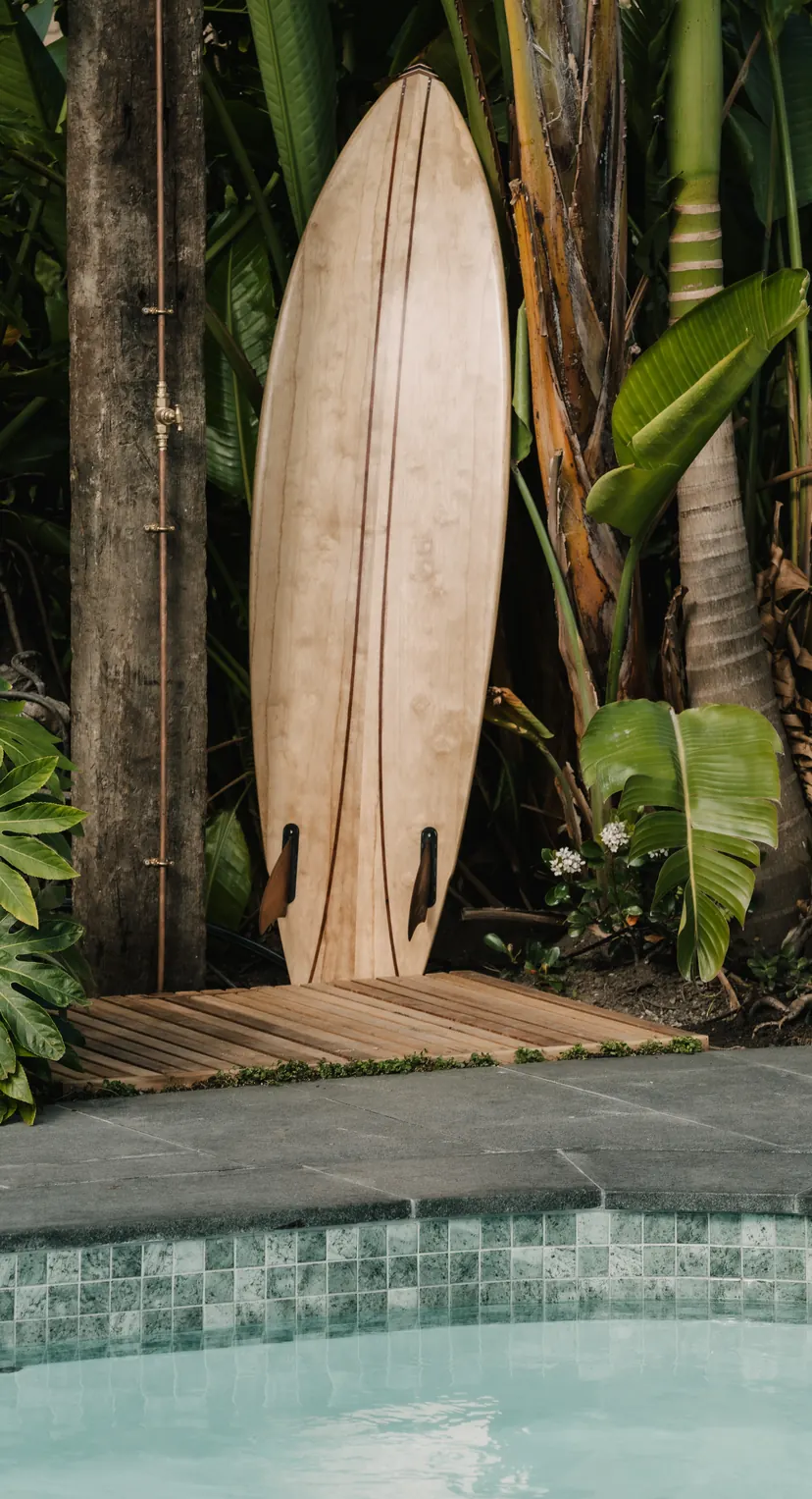 A timber surfboard standing among the palms at the pool's edge, the water below
